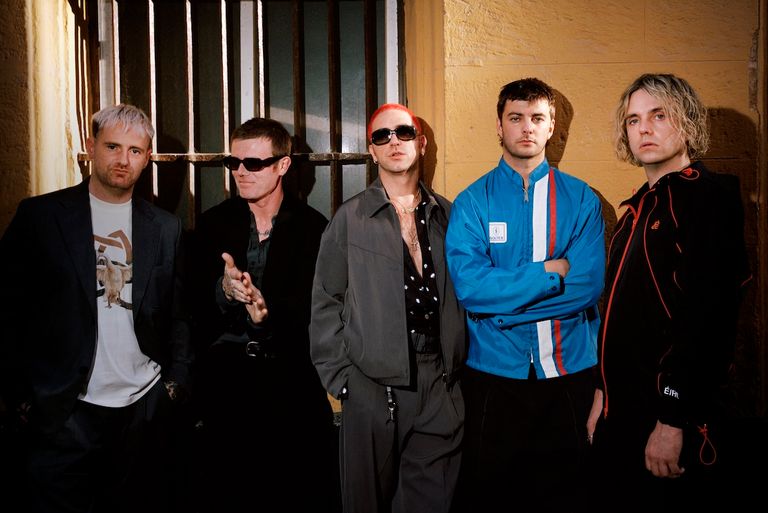 Post-punk band Fontaines D.C. standing together in dark streetwear and jackets against a yellow wall, photo by Ewen Spencer.